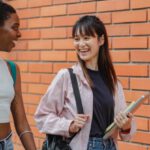 Campus Activities - Cheerful multiracial female students wearing casual clothes carrying backpack and notebooks near college brick wall while discussing plans and looking at each other with smiles