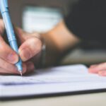 Scholarship Essay - Person Holding Blue Ballpoint Pen Writing in Notebook
