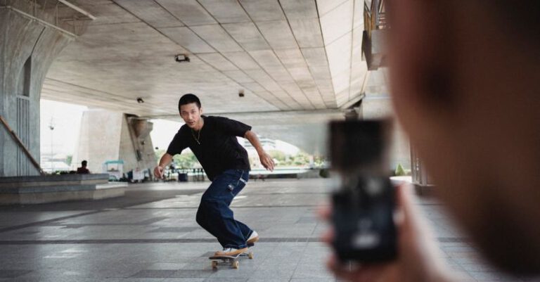 Memory Tricks - Full body of sportive man riding on skateboard under bridge and looking at camera of smartphone during training in street