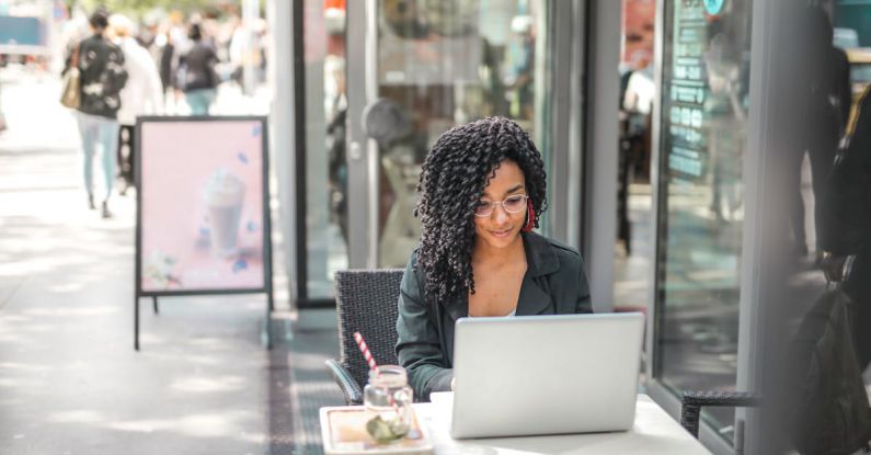 Online Degree Programs - High angle of pensive African American female freelancer in glasses and casual clothes focusing on screen and interacting with netbook while sitting at table with glass of yummy drink on cafe terrace in sunny day